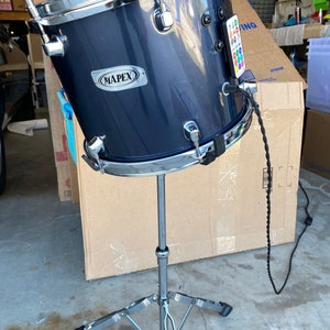 May include: A blue drum with a chrome stand and a Mapex logo. The drum has a black cord attached to it.