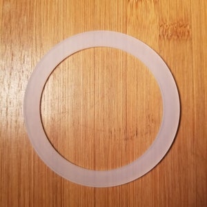 Op de afbeelding: Een doorschijnende, crèmekleurige, cirkelvormige pakking. De pakking is gemaakt van een flexibel materiaal en staat op een houten achtergrond. De pakking heeft een ringvorm met een consistente breedte.