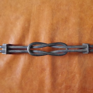 May include: A metal object with a figure-eight knot design. It has two rectangular ends, each with three prongs. The metal is dark grey, set against a textured, brown background. The object is likely handmade.