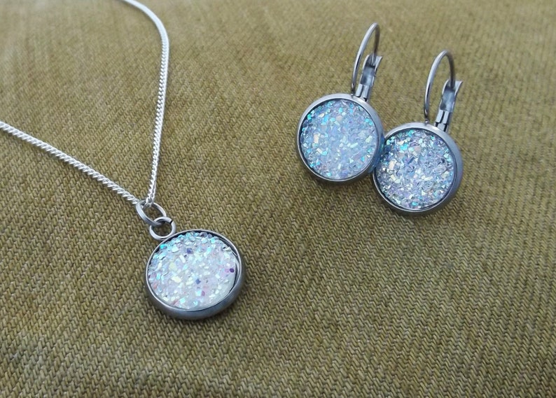 May include: A silver chain necklace and matching earrings with a round pendant and earrings featuring a white glitter design.