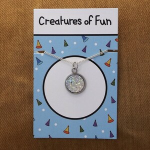 May include: A silver necklace with a round, white, sparkly pendant. The pendant is on a card with the text "Creatures of Fun" and a blue background with white dots and colorful party hats.