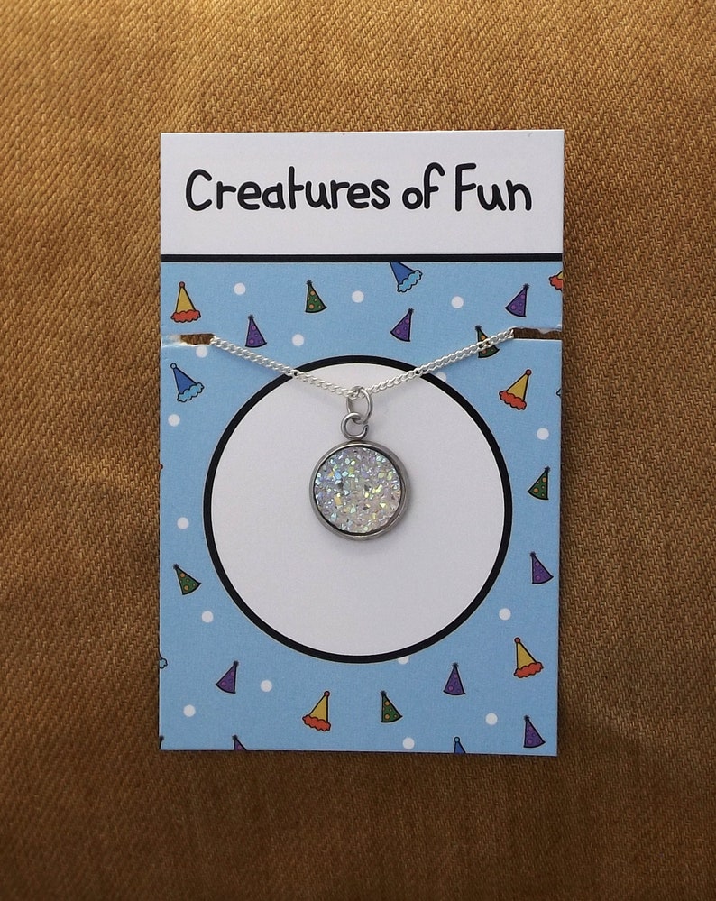 May include: A silver necklace with a round, white, sparkly pendant. The pendant is on a card with the text "Creatures of Fun" and a blue background with white dots and colorful party hats.