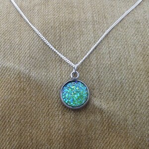 May include: A silver chain necklace with a round pendant featuring a green and blue glitter-filled resin design.