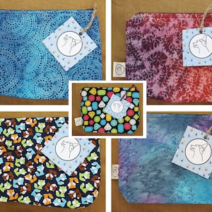 Pode incluir: Cinco bolsas de zíper de tecido diferentes com um cão usando um chapéu de festa em uma etiqueta. As bolsas são de vários padrões, incluindo azul com pontos brancos, vermelho com flores roxas, azul com raposas brancas e laranja, azul com tie-dye branco e laranja, e preto com joias coloridas.