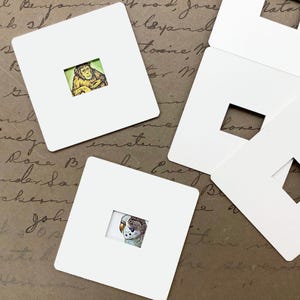 May include: Six white square frames with a cut-out window in the center. The frames are on a brown surface with handwritten text. Two of the frames have images inside: a chimpanzee and a parakeet.