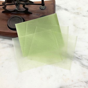 May include: Four translucent green sheets of wax paper, stacked on top of each other, on a white marble surface.