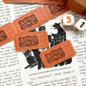Puede incluir: Primer plano de tres boletos de papel naranja vintage con el texto "ADMIT ONE" impreso en ellos. Los boletos est&aacute;n impresos en tinta negra y tienen un n&uacute;mero de serie impreso en cada uno. Los boletos est&aacute;n sobre un libro con ilustraciones en blanco y negro de gatos.