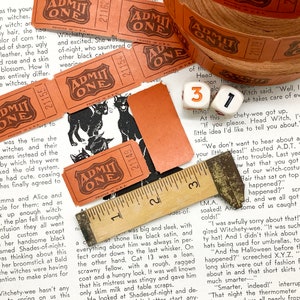 Puede incluir: Una colecci&oacute;n de boletos vintage "ADMIT ONE" impresos en naranja y marr&oacute;n con texto negro. Los boletos presentan una ilustraci&oacute;n de un gato negro. Los boletos est&aacute;n dispuestos en una p&aacute;gina de libro con una regla y dados.