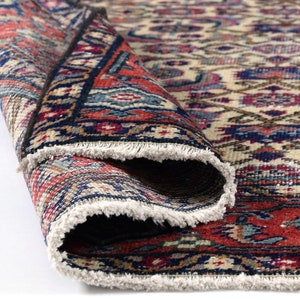 Puede incluir: Primer plano de una alfombra enrollada con un patrón detallado e intrincado en tonos rojos, azules y crema. La alfombra tiene un borde con flecos.