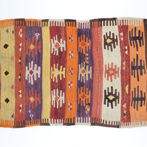 K&ouml;nnte beinhalten: Ein bunter Vintage-Teppich mit einem geometrischen Muster in Rot-, Orange-, Gelb-, Braun- und Violettt&ouml;nen. Der Teppich hat einen Fransenrand und ist mit einem traditionellen Muster gewebt.