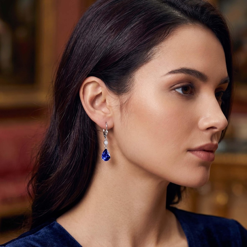 May include: Elegant silver earrings with a teardrop-shaped, deep blue gemstone and a small, round, clear stone. The earrings are dangling, with a lever-back closure. The woman in the image is wearing a dark blue velvet top.