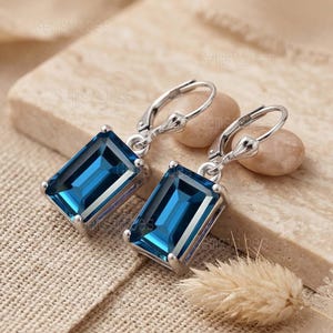 May include: A pair of silver-toned leverback earrings with rectangular, deep blue gemstones. The gemstones are set in a silver frame, and the earrings are displayed on a textured, neutral-toned surface. The earrings have a polished finish.