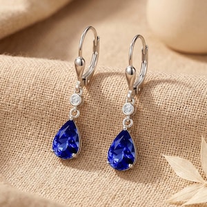May include: A pair of silver-toned leverback earrings. Each earring features a teardrop-shaped, faceted, deep blue gemstone suspended below a small, round, clear stone. The earrings are displayed on a neutral-toned fabric.