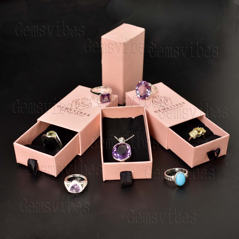 May include: Pink jewelry boxes with black interiors display rings and a pendant. Rings feature various gemstones, including amethyst and turquoise. The boxes are arranged on a dark surface, showcasing the jewelry.