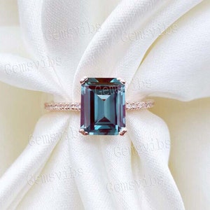 May include: Rose gold ring with an emerald-cut, teal gemstone. The band has a textured pattern. The ring is set against white fabric, creating a luxurious aesthetic. The gemstone's dimensions are not visible.