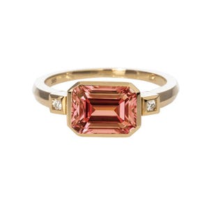 May include: A gold ring featuring a large, rectangular, pink gemstone. Two smaller diamonds flank the main stone. The ring has a simple, elegant design, perfect for everyday wear or special occasions. The ring is a beautiful piece of jewelry.