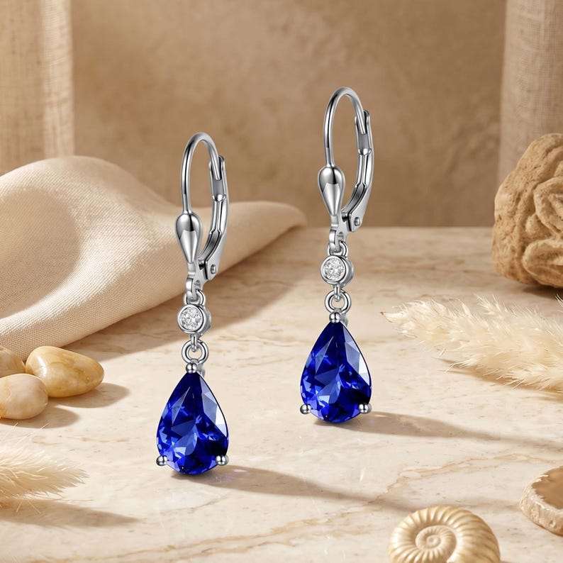 May include: A pair of silver-toned drop earrings. Each earring features a teardrop-shaped, deep blue gemstone suspended below a small, round, clear stone. The earrings have a lever-back closure.