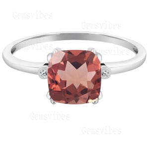 May include: A silver ring featuring a large, square-cut, reddish-brown gemstone as the centerpiece. Small, round, clear accent stones are set on either side of the main stone. The ring has a simple, elegant design.