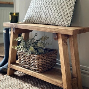 May include: A rustic wooden bench with a lower shelf, featuring a patterned cushion and a wicker basket filled with greenery. The bench is stained a warm brown colour and has a simple, farmhouse style.