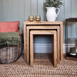 May include: Three wooden nesting tables with a natural wood finish. The tables are stacked on top of each other and have a simple, rustic design. A white jug with green foliage sits on the top table, and two clear glass candle holders are on the table below.