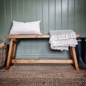 May include: A rustic wooden bench with a white throw blanket and a pillow. The bench is in front of a green wall with wood paneling.