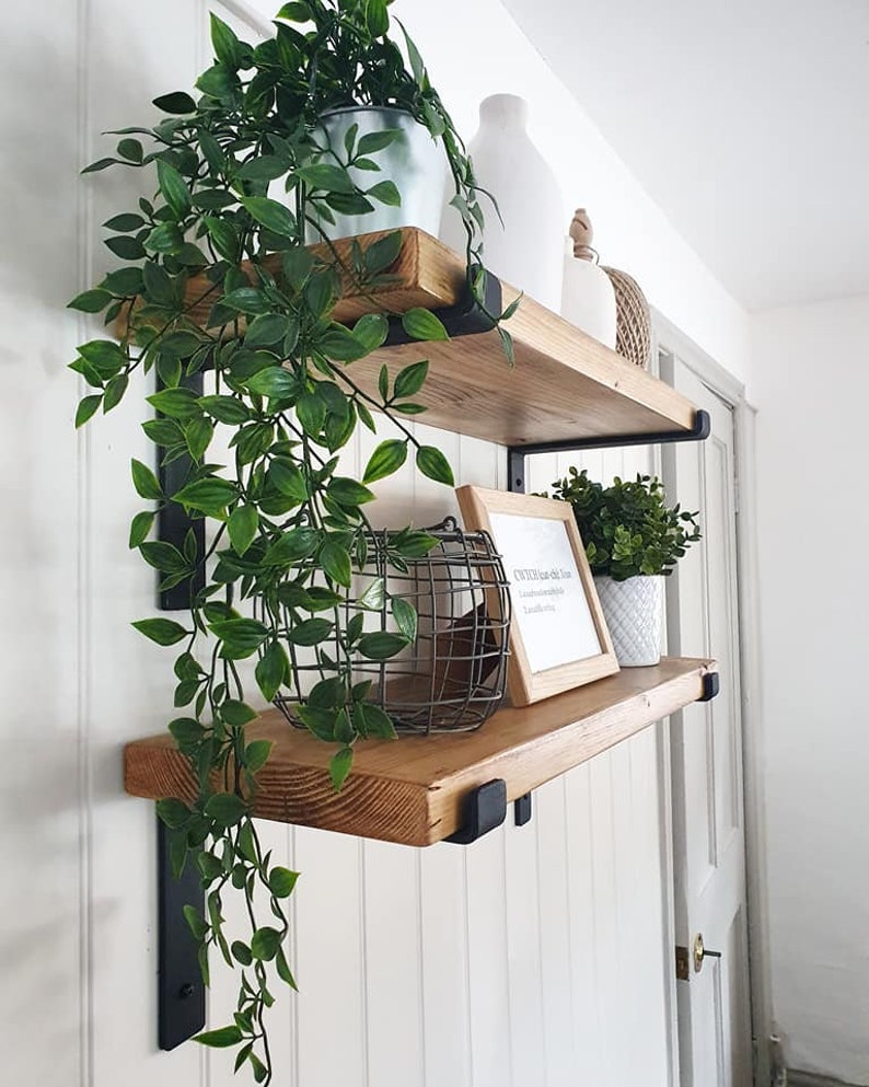 May include: Two wooden floating shelves with black metal brackets on a white wall. The shelves display decorative items: potted plants, a wire basket, a framed print, and white vases. Trailing green plants cascade from the top shelf, adding a natural touch.
