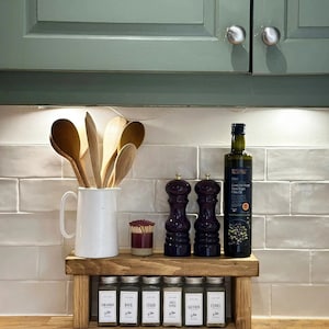 May include: A wooden spice rack with a shelf for storing spices and bottles. The rack has a white ceramic pitcher with wooden spoons, a bottle of olive oil, and two salt and pepper shakers. The rack is on a wooden countertop with white tile backsplash.