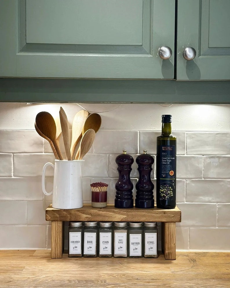 May include: A wooden spice rack with a shelf for storing spices and bottles. The rack has a white ceramic pitcher with wooden spoons, a bottle of olive oil, and two salt and pepper shakers. The rack is on a wooden countertop with white tile backsplash.