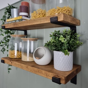 May include: Two wooden shelves with black metal brackets fixed to a pale green wall. The shelves display glass jars filled with pasta, rice, and other items, plus decorative plants and candles. The shelves are made of stained wood.