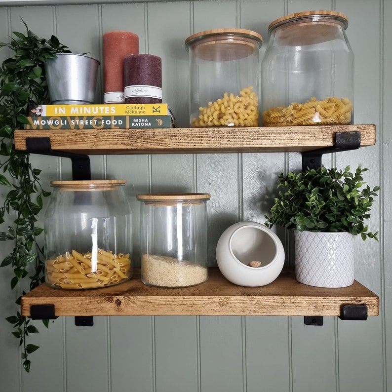 May include: Two rustic wooden shelves with black metal brackets are mounted on a green wall. The shelves display glass jars with pasta, rice, and decorative items. Books, candles, and plants add to the decor. The shelves are a stylish storage solution.