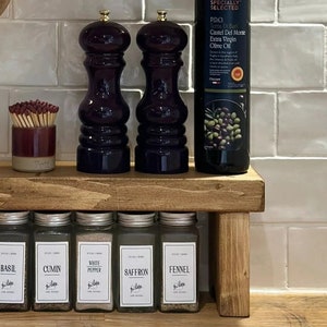 May include: A wooden spice rack with five glass jars filled with spices labeled "Basil", "Cumin", "White Pepper", "Saffron", and "Fennel". Two black pepper grinders and a bottle of olive oil are on the shelf above the spice rack.