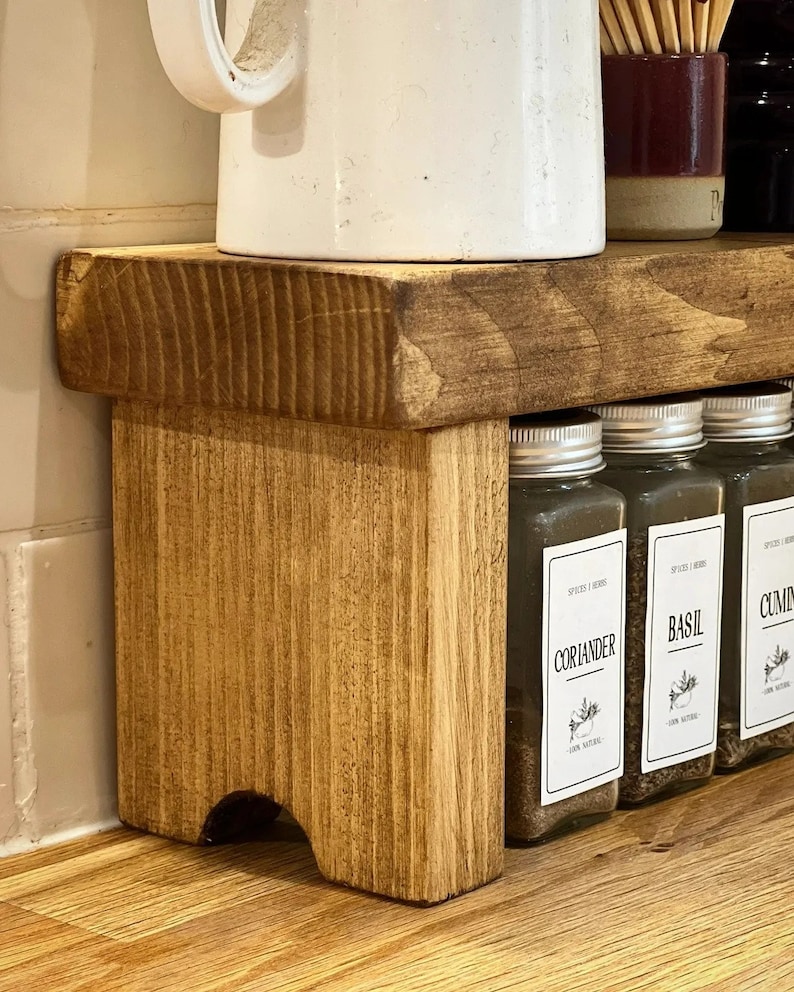 May include: A wooden spice rack with four glass jars filled with spices. The jars have labels that read "Coriander", "Basil", "Cumin", and "Oregano". The rack is stained a dark brown color and has a rustic look.