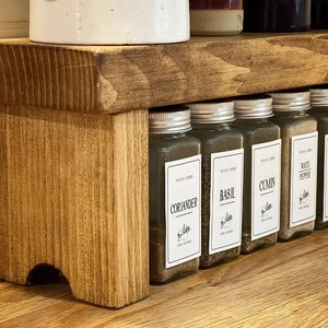 May include: A wooden spice rack with five glass jars filled with spices. The jars have labels that read "Cumin", "White Pepper", "Basil", "Coriander", and "Oregano".