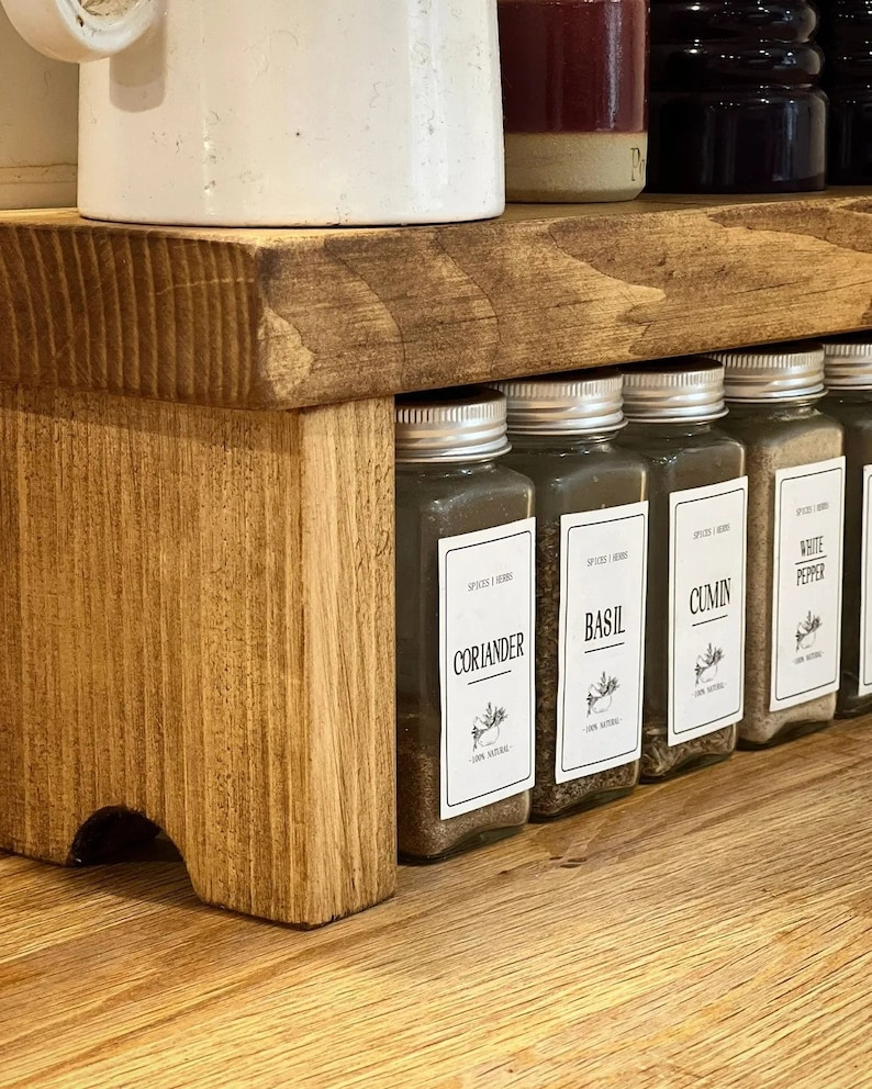 May include: A wooden spice rack with five glass jars filled with spices. The jars have labels that read "Cumin", "White Pepper", "Basil", "Coriander", and "Oregano".