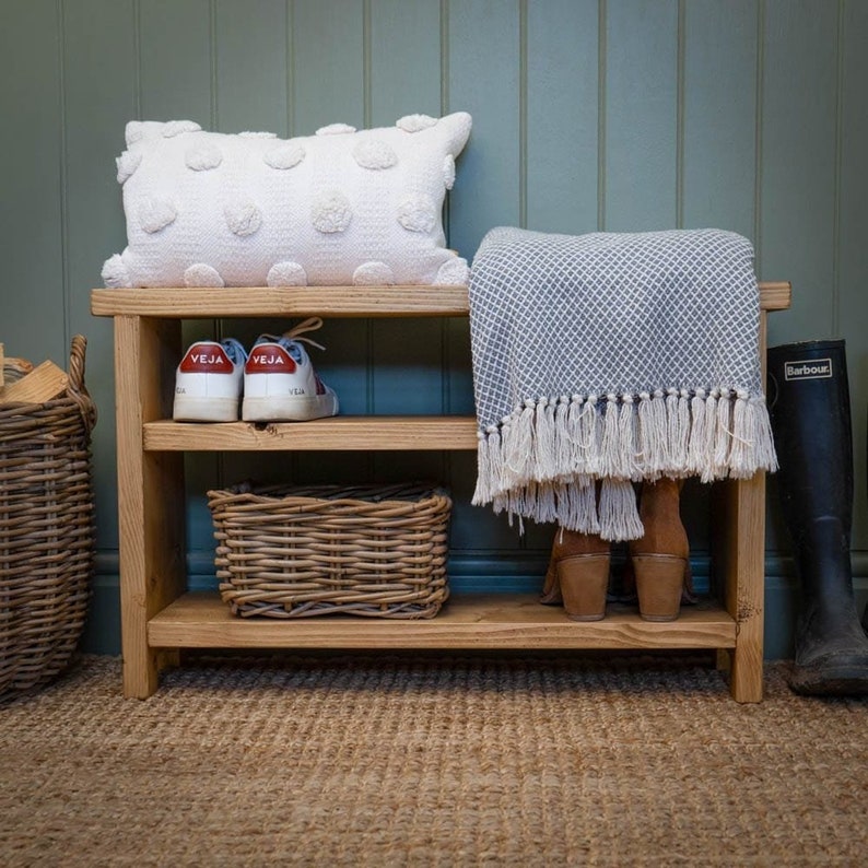 May include: A wooden shoe rack with two shelves, a white throw pillow, a gray throw blanket, a wicker basket, and a pair of white sneakers with red accents. The shoe rack is against a green wall.