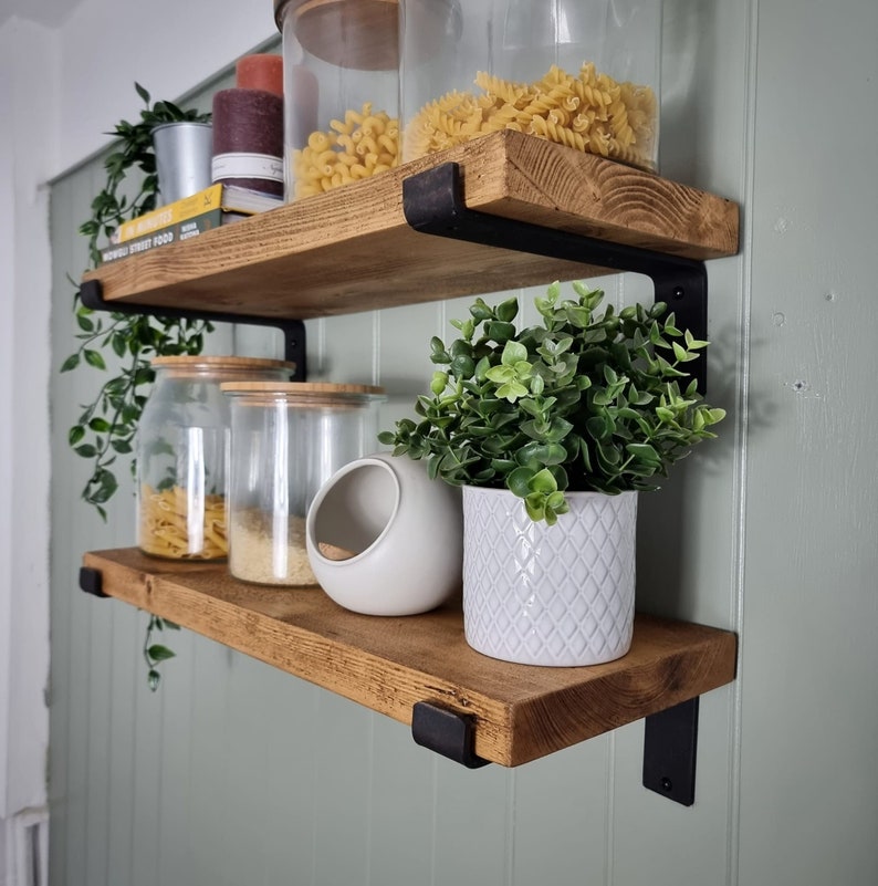 May include: Two wooden shelves with black metal brackets fixed to a pale green wall. The shelves display glass jars of pasta, a white ceramic salt cellar, and a potted plant. Other items include a candle and books.