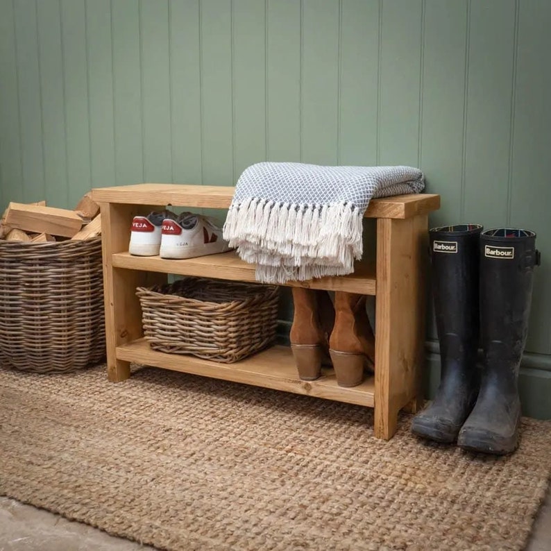 May include: A wooden shoe rack with two shelves and a top shelf for storage. The rack is made of natural wood and has a rustic look. It is filled with shoes, boots, and a blanket.