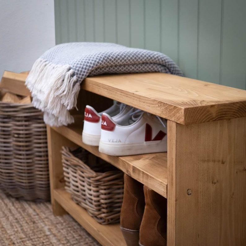 May include: A wooden shoe rack with two shelves and a bench top. The rack is made of natural wood and has a rustic look. There are white sneakers with red accents on the top shelf and brown boots on the bottom shelf. A gray and white blanket is folded on the bench top.