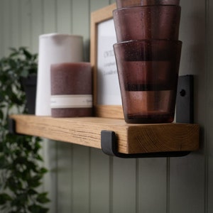 May include: A wooden shelf with black metal brackets, featuring a white jug, a brown candle, and a stack of four clear glass tumblers with a purple hue.