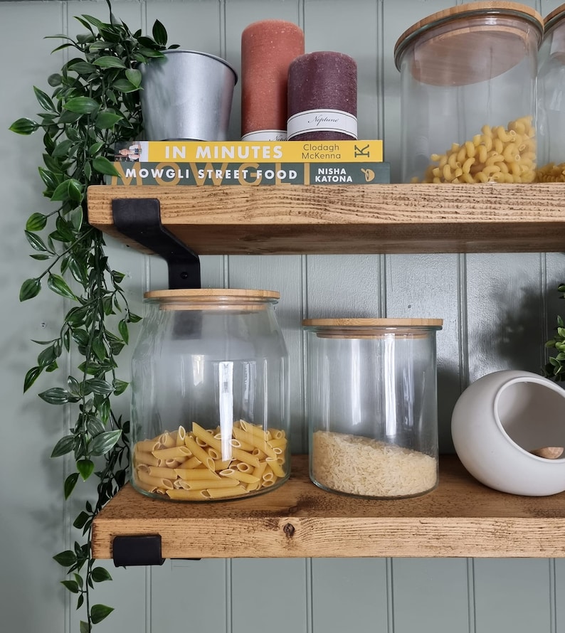 May include: A wooden shelf unit features glass storage jars with bamboo lids, containing pasta and rice. Candles, a metal bucket, and cookbooks are also present. Trailing greenery adds a natural element to the kitchen storage. The shelf is supported by black brackets.