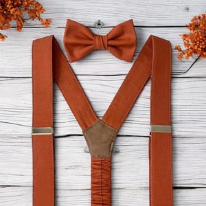 May include: A set of burnt orange bow tie and braces. The braces have silver-coloured metal clips and a brown leather-like patch at the back. The bow tie is the same colour and fabric. The set is displayed on a white wooden surface.