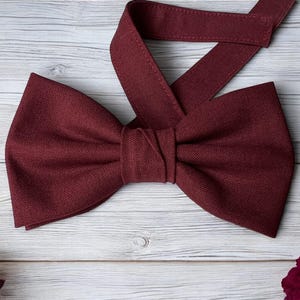 May include: A burgundy bow tie made from textured fabric, with a matching adjustable neck strap. The bow tie is on a light wood-grain surface, with burgundy roses in the corners. A formal accessory.