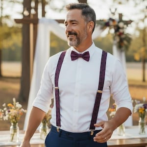 May include: A man in a white dress shirt, navy blue pants, and a burgundy bow tie and suspenders. The suspenders have a subtle pattern. The background includes a wooden structure and outdoor setting, suggesting a wedding or formal event.