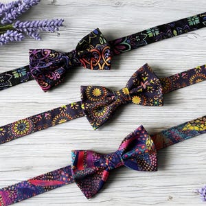 May include: Three patterned bow ties in shades of purple, yellow, and red, with matching neck straps. The bow ties are displayed on a light wooden surface, with sprigs of lavender. Each strap is adjustable.