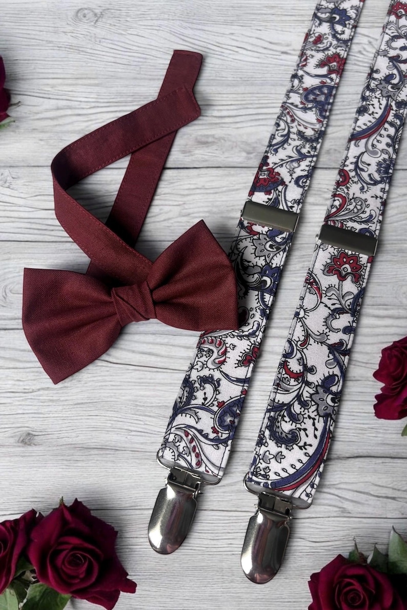 May include: A burgundy bow tie and patterned suspenders with silver clips. The suspenders feature a white background with a paisley design in red, blue, and gray. The bow tie and suspenders are displayed on a light wood surface.