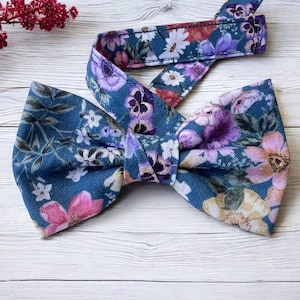May include: A floral bow tie and matching neck strap. The teal fabric is patterned with pink, purple, and white flowers. The accessory is displayed on a white wooden surface, with red floral decorations.