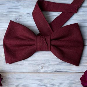 May include: A burgundy bow tie with a matching adjustable neck strap. The bow tie is made of a textured fabric and is centered on a light wood grain surface. Dark red roses are scattered around the bow tie.