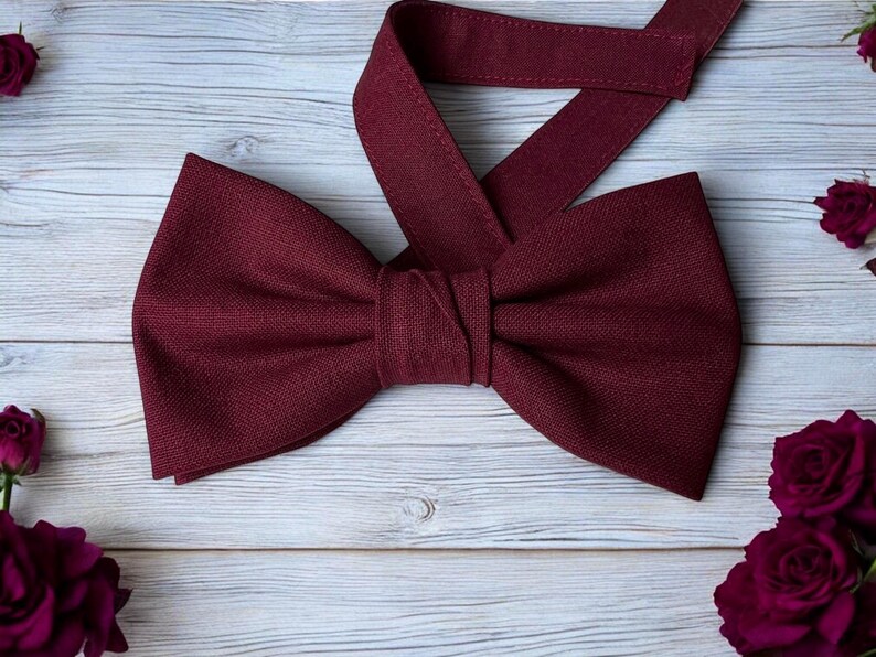May include: A burgundy bow tie with a matching adjustable neck strap. The bow tie is made of a textured fabric and is centered on a light wood grain surface. Dark red roses are scattered around the bow tie.