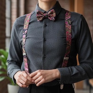 May include: A gray button-down shirt paired with black pants, suspenders, and a bow tie. The suspenders and bow tie feature a paisley pattern with pink, green, and black accents. The outfit is a stylish ensemble.