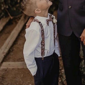 May include: A young boy in a white button-down shirt, navy pants, and floral suspenders and bow tie. He is looking up with a smile. The boy is standing next to a person wearing a dark suit.